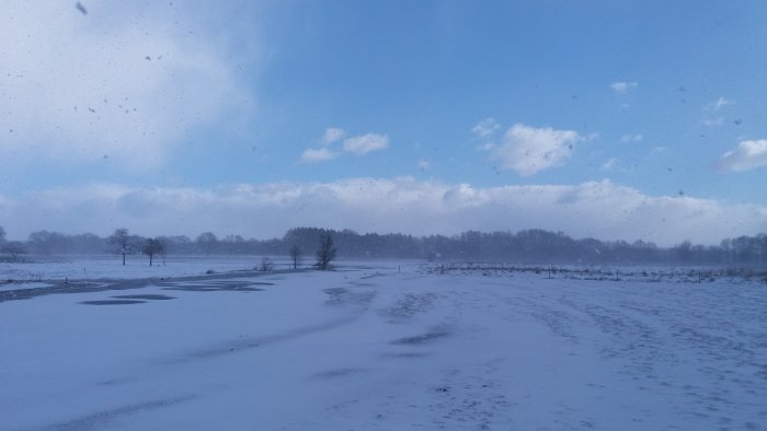 Växlande väder över snötäckt fält med blå himmel och drivande snöflingor.