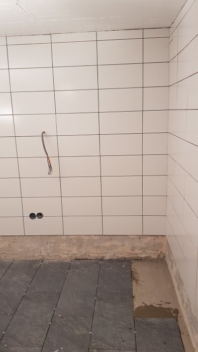 Vägg med nytt vitt kakel runt avslutad installation och delvis klinklat golv med svarta plattor.