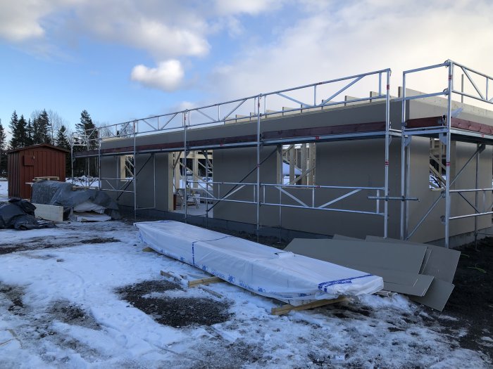 Garage i byggfas med ställningar och osatt panel, material på marken, snö och blå himmel i bakgrunden.