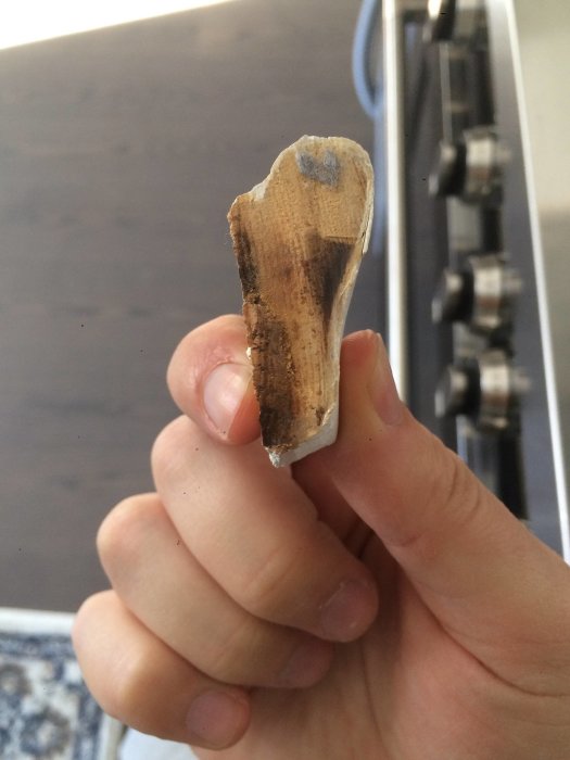 En hand håller en avbruten bit av trälist med synlig profil nära kameran, mot bakgrund av köksutrustning.