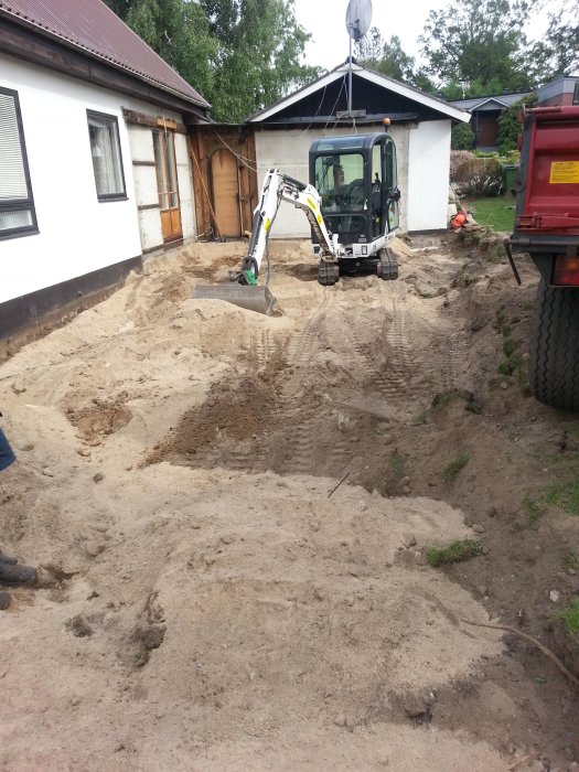 Grävmaskin i arbete med att gräva i en trädgård bredvid ett hus, grushög och spår i sanden syns.