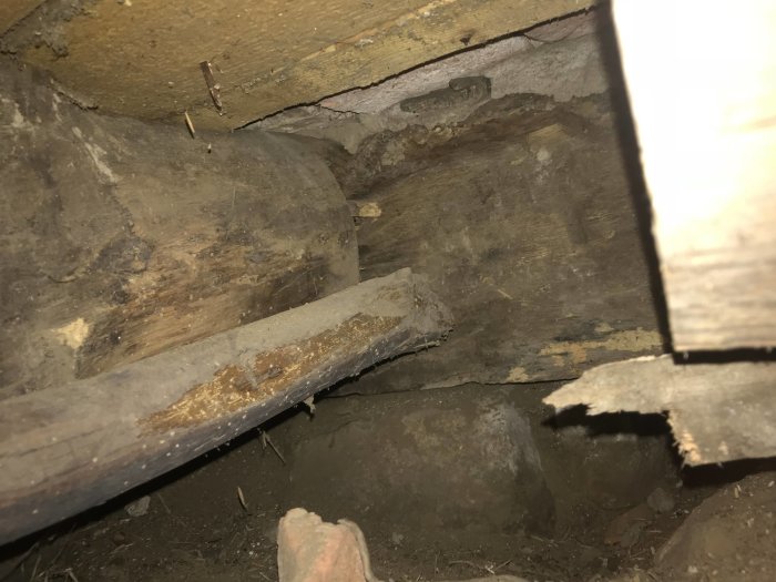 Mörkt kryputrymme under en byggnad med synliga träbjälkar och tegelstenar, tecken på slitage och förfall.