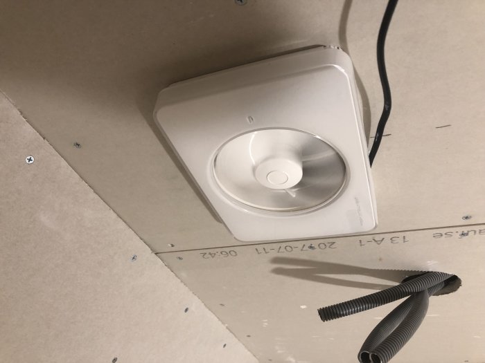 Nyinstallerad vit badrumsfläkt på gipsvägg med synlig elektrisk kabel och ventilationsslang.