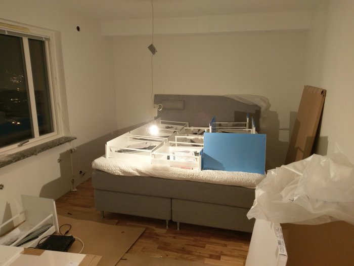 Sovrum med pågående renovering, säng täckt av lådor och förpackningar, omonterade skåpsdelar på golvet.