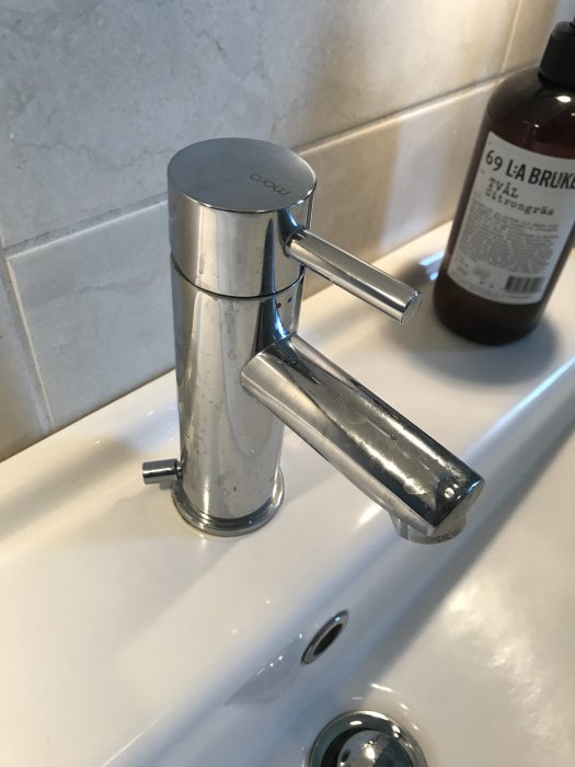 Kromad blandare med två handtag monterad på vit tvättställ med tvålvask i bakgrunden.