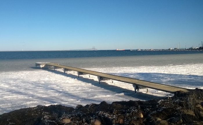 Bild på en enkel träbrygga som sträcker sig ut över ett snötäckt sund med en bro i bakgrunden.