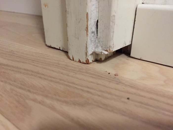 Skadade vita golvlister bredvid ett laminatgolv som inte matchar runt en dörrfodring.