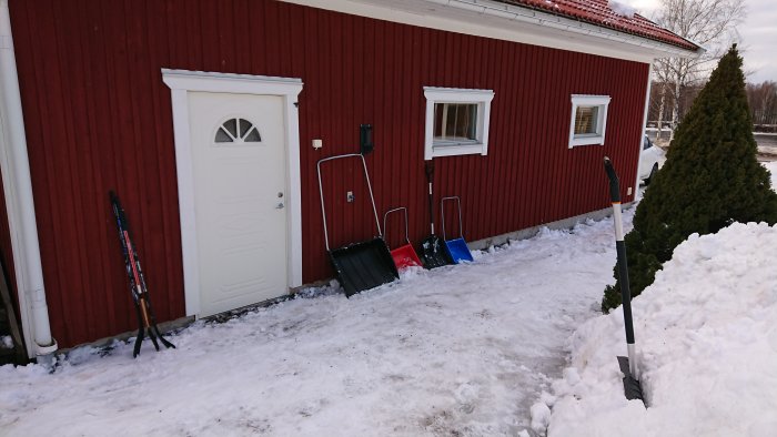 Ett rött hus med tre snöskyfflar lutade mot väggen och skidstavar, snö röjd på uppfart.