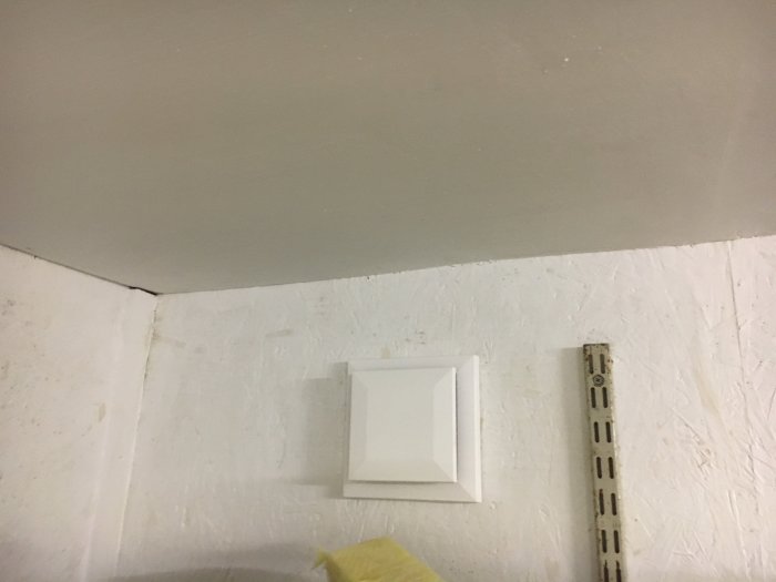 Ventilationslock på en vitmålad vägg i ett garage med en fuktmätare som visar hög luftfuktighet.