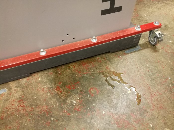 Oljeutsläpp på betonggolv bredvid en röd maskinpelare med hjul legs
