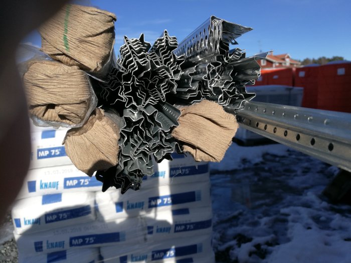 Byggmaterial med träreglar och metallprofiler framför säckar av Knauf MP 75 på en byggarbetsplats.