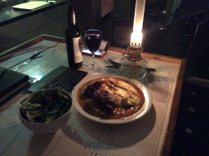 Middagsscenario med skomakarlåda, sallad, rödvin och levande ljus på ett bord vid datorskrivbordet.