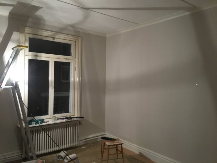 Renoveringsprojekt med nytapetserad vägg nära fönster och en stege, överblivet tapet och verktyg på golvet.