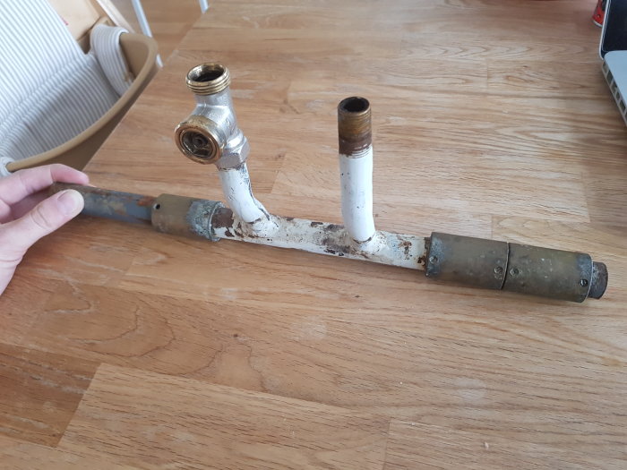 Hand håller rörkonstruktion med bypass och kopplingar från ett demonterat element, mot trägolv bakgrund.