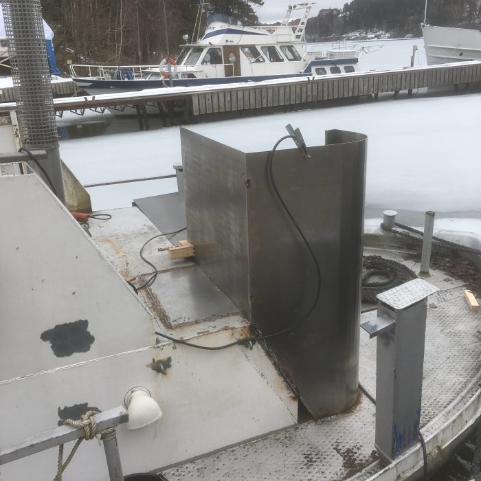 Metallkonstruktion på en båtbrygga med snötäckt isigt vatten och båtar i bakgrunden.