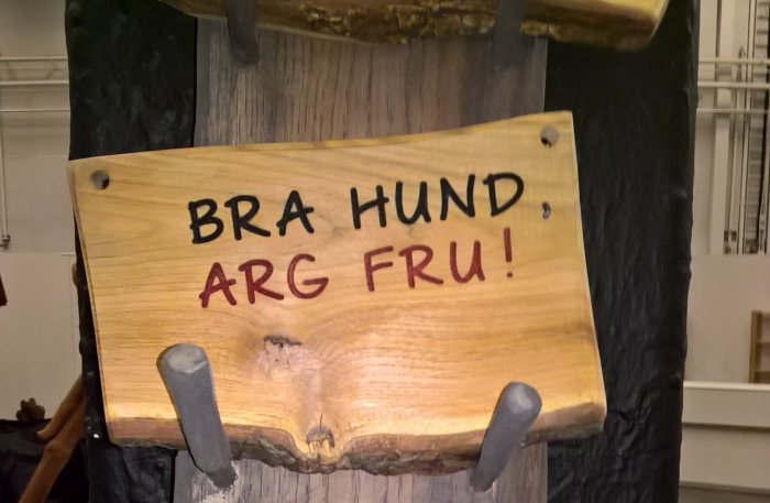 Skylt på träplank med texten "BRA HUND, ARG FRU!" och två metallkrokar.