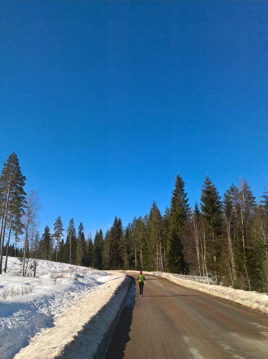 En person joggar på en solig väg omgiven av snötäckta träd och klarblå himmel.