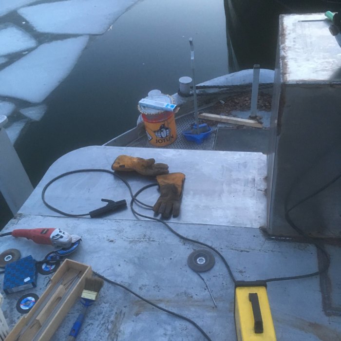 Byggarbetsplats vid skymning med verktyg, svetshandskar och en burk målarfärg på en plåtyta bredvid isbelagt vatten.