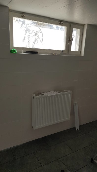 Nyinstallerat vitt element under ett fönster i en tvättstuga med gråa klinkergolv och kakelväggar.