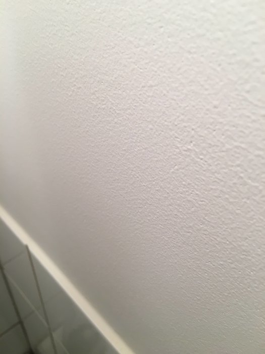 Nymålad vit vägg med synlig textur och en del av ett vitt kakel i nedre kanten.