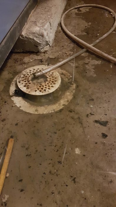 Vattenansamling runt en golvbrunn i en källare med synlig betong och en gammal rostig pannplåt i bakgrunden.
