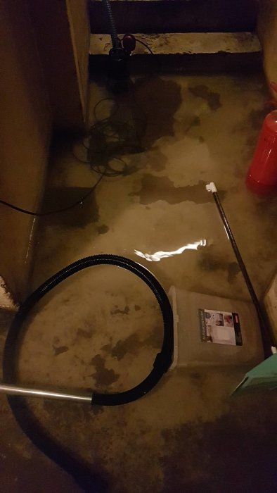 Källargolv med vattensamlingar nära en pump och slangar, indikerar problem med dräneringen.