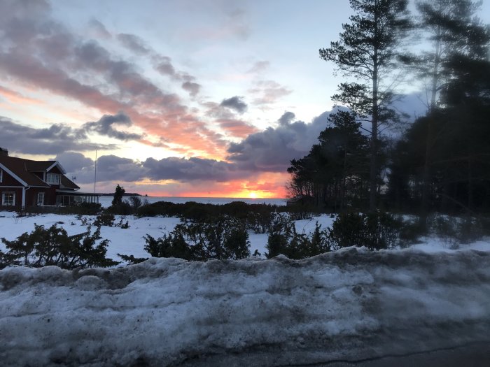 Solnedgång med dramatiska moln över snötäckt landskap och hus, sedd från en väg.