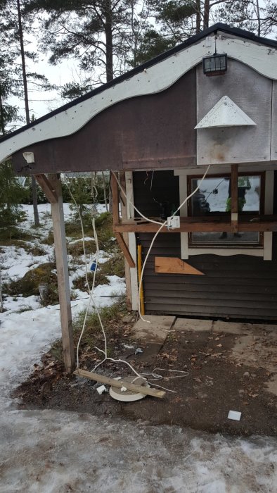 Friggebod under renovering med synligt träverk och byggavfall på marken, delvis snötäckt mark.