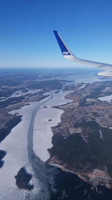 Flygplansvinge över snötäckt landskap och isbelagd flod sedd från ovan.