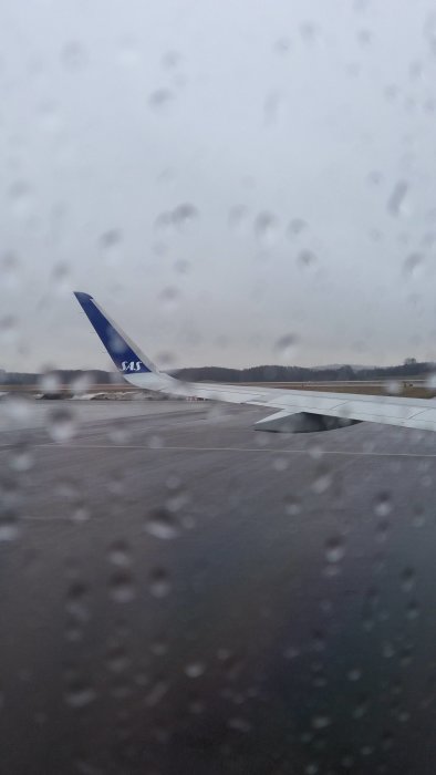 Flygplansvinge från SAS-flyg med regndroppar på fönstret och grå himmel i bakgrunden.