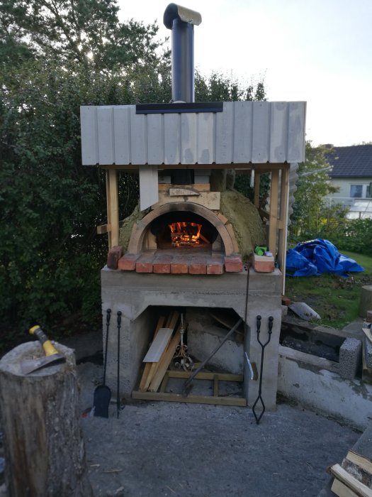 Hembyggd uteugn med eld inuti och vedförvaring under, omgiven av verktyg och natur.