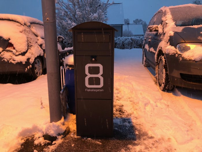 Ny brevlåda med nummer 8 och namnet 'Fiskalsgatan' på en snöig gata vid skymningen.