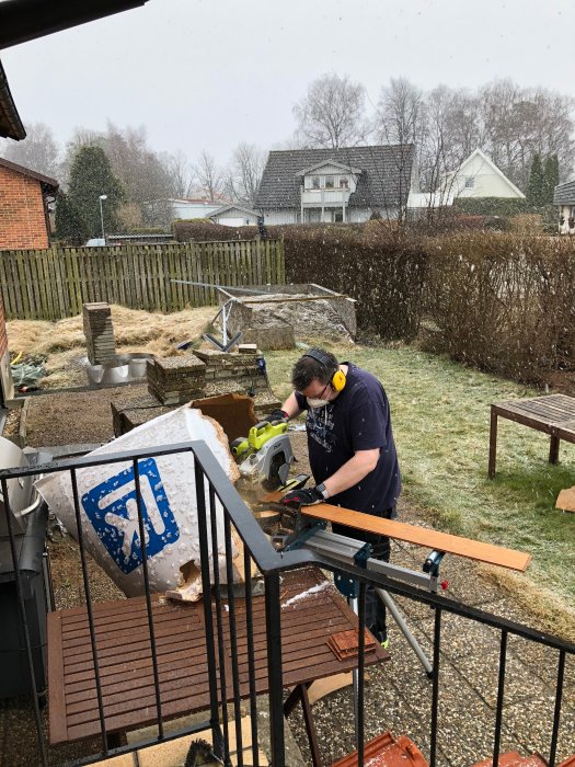 Person sågar golvplank på farstukvist med snöfall i bakgrunden.