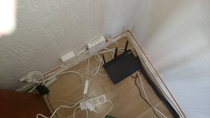 Rörig hörna med modem, sladdar och grenuttag på ett trädgolv mot en vit vägg.