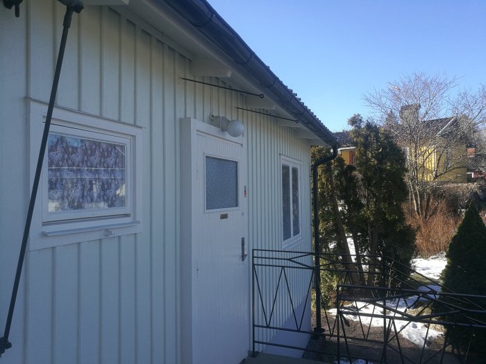 Exteriör av ett vitt hus med en dörr och fönster, snö på marken och gröna buskar, i Småland.