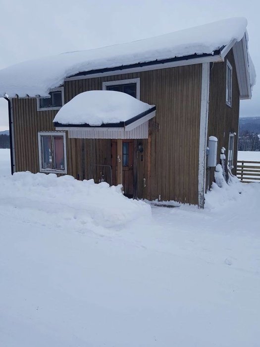 Trähus delvis täckt av snö med tjocka snödrivor framför och på taket.
