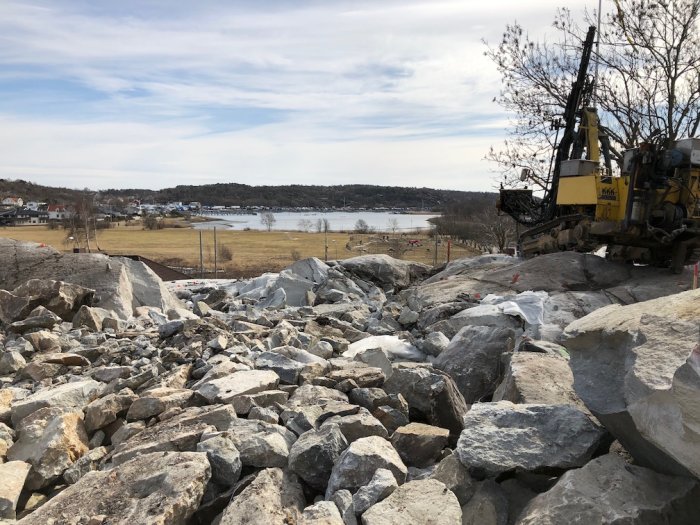 Borrningsmaskin på klippa med sprängda stenblock framför, utsikt över landskap och vatten.