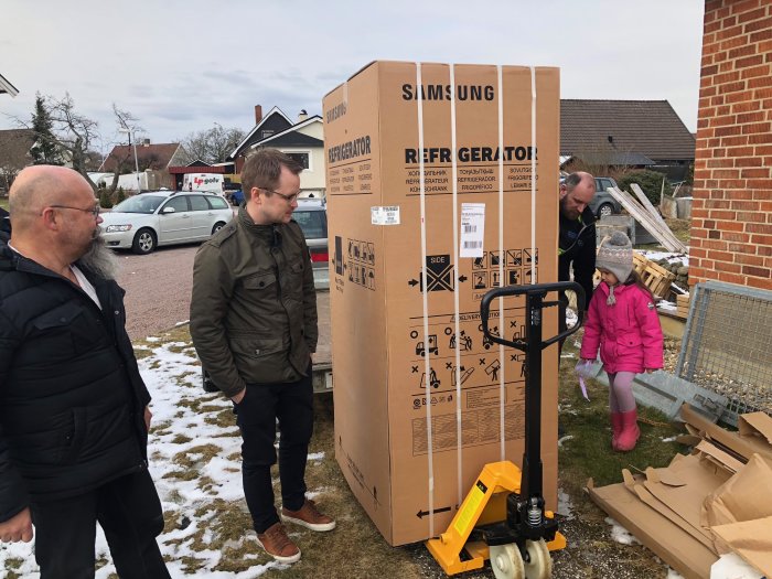 Tre personer och en stor kartong med kylskåp utomhus, nära en pallbock med snö på marken.