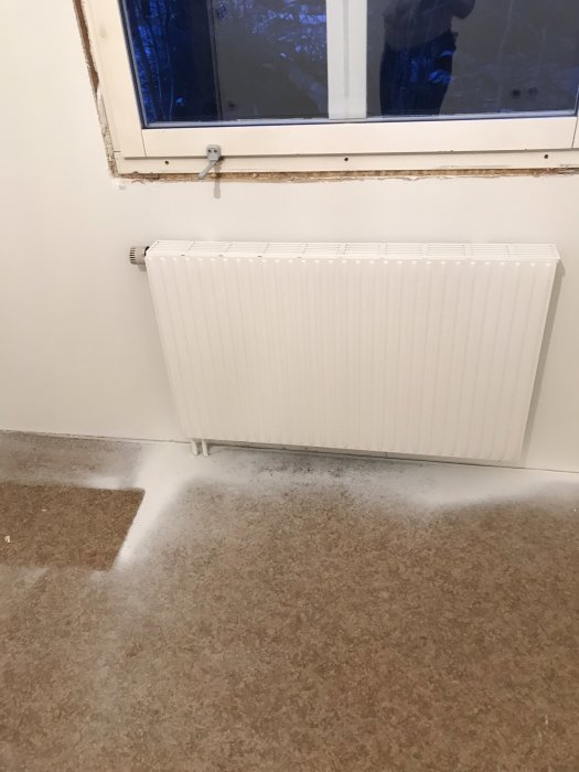 Radiator under ett fönster i ett orenoverat rum med synliga rör och avskalat område runt fönstret.