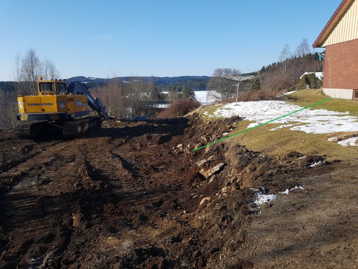 Grävmaskin vid arbete med sluttning från hus till garage, ojämn gräsmatta med snörester.