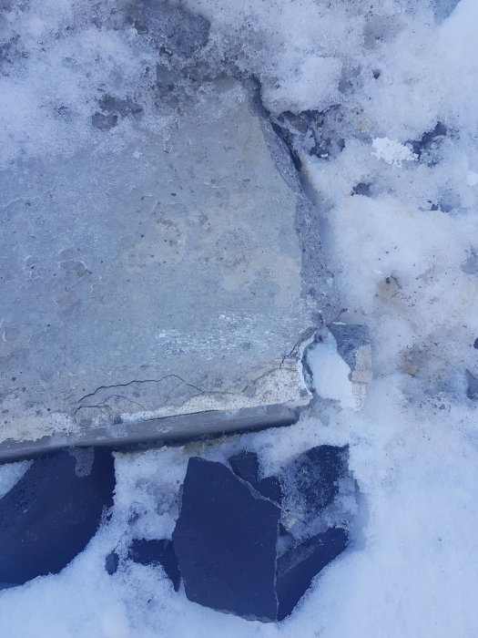 Skadad kant på garageplatta med sprickor och brutna betongbitar i snö.