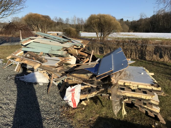 Hög med rivet byggavfall av trä och isolering på en grusplan med snö och landskap i bakgrunden.