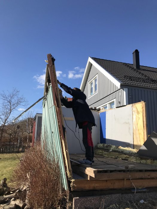 Person avlägsnar delar av ett rivet förråd bredvid ett hus.