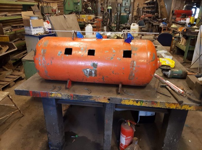 Gammal sliten orange kompressortank med utskurna öppningar på ett arbetsbord i en verkstad.