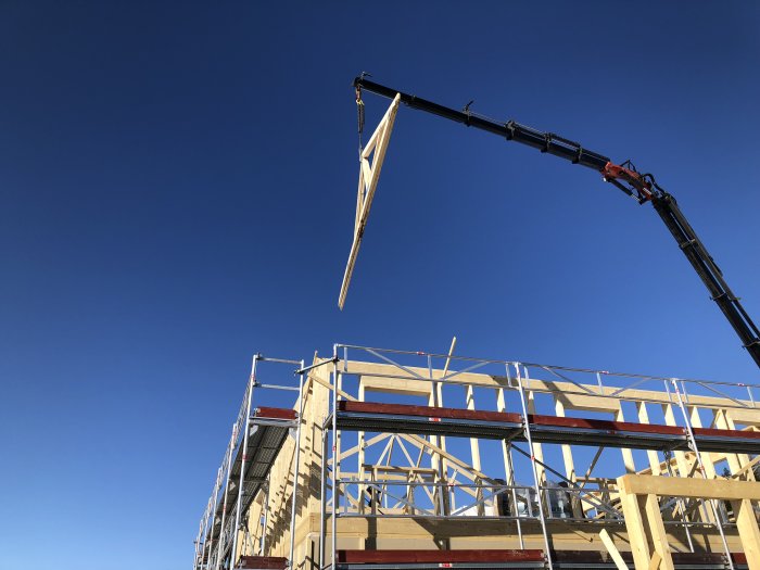 Kranbil lyfter takstol på en byggnadsstomme mot klarblå himmel.