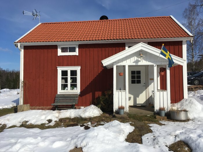 Röd stuga med vita knutar och snötäckt mark, veranda till höger, svensk flagga, och soligt väder.