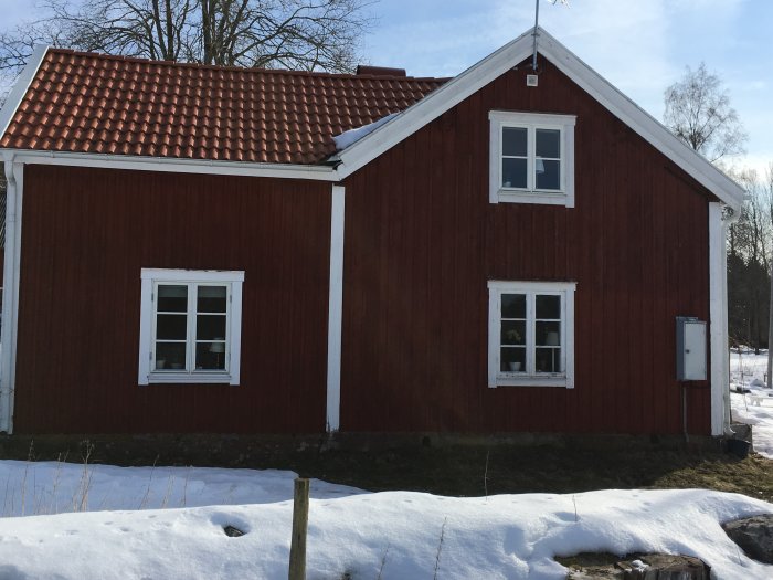 Rödmålat hus med snötäckt mark och gavel till höger nära verandan under en klar himmel.