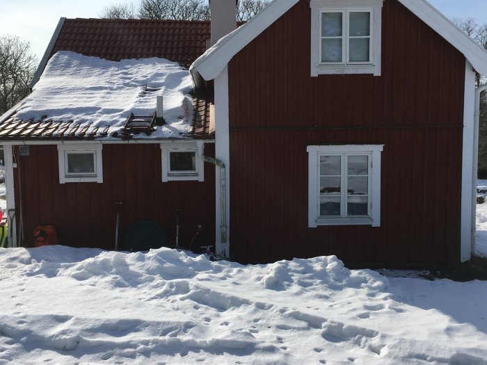 Röd trävilla med snötäckt tak och veranda under vintersol, potentiell gavelutbyggnad till höger.