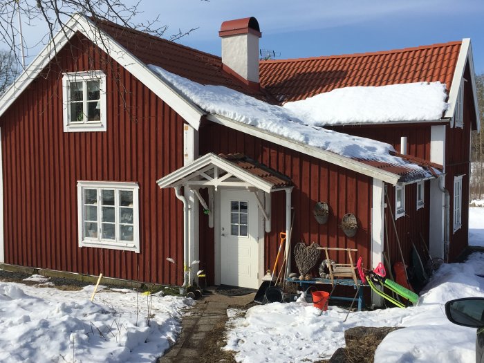 Rödmålat hus med snö på taket, vit dörr, fönster och veranda, med diverse föremål framför.