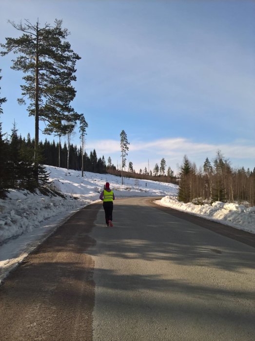 Person i reflexväst joggar på en väg omgiven av snötäckta träd och blå himmel.